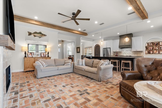 living room featuring a tiled fireplace, ceiling fan, ornamental molding, and beamed ceiling