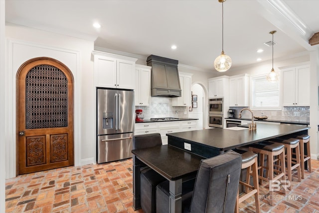 kitchen with premium range hood, appliances with stainless steel finishes, decorative light fixtures, white cabinets, and a kitchen island with sink