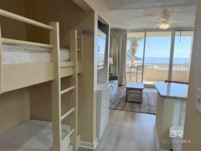 bedroom featuring light wood-type flooring, a textured ceiling, ceiling fan, expansive windows, and a water view