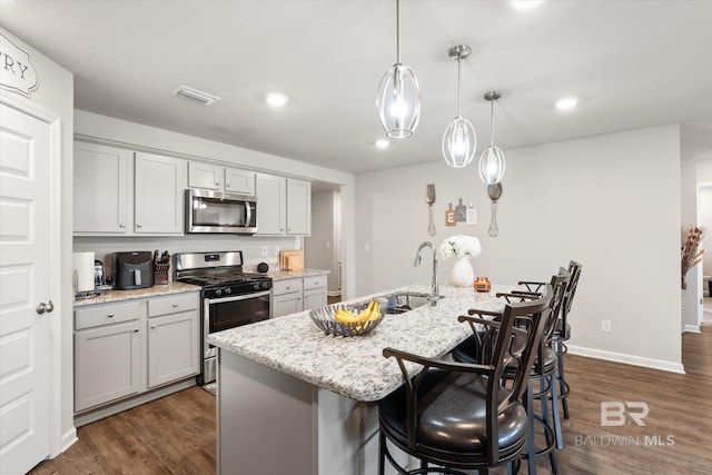 kitchen with light stone countertops, dark wood-type flooring, appliances with stainless steel finishes, decorative light fixtures, and a center island with sink