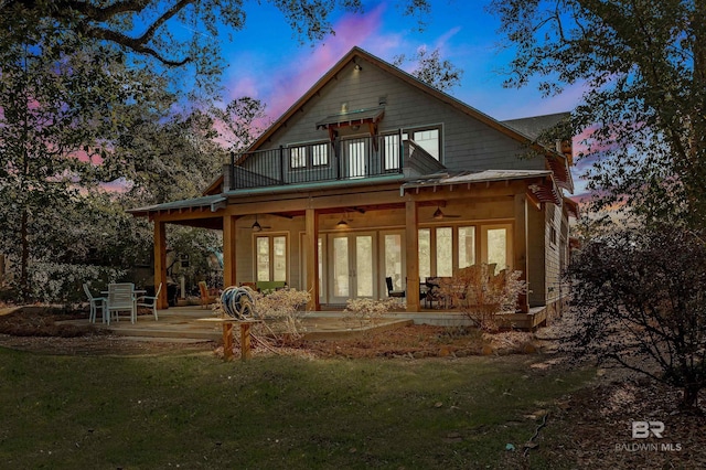 back of house at dusk featuring a balcony, a ceiling fan, french doors, a lawn, and a patio area