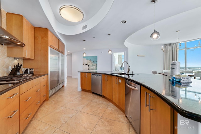 kitchen with a sink, backsplash, dark stone counters, appliances with stainless steel finishes, and wall chimney exhaust hood