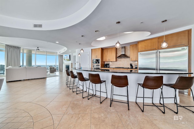 kitchen with dark countertops, visible vents, stainless steel built in refrigerator, a breakfast bar area, and wall chimney exhaust hood