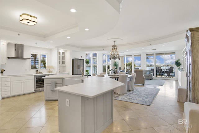 kitchen featuring wine cooler, an island with sink, a raised ceiling, wall chimney exhaust hood, and a sink