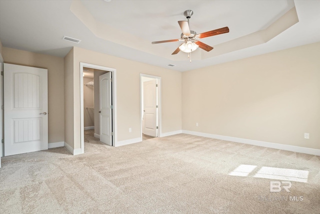 unfurnished bedroom featuring light carpet, ensuite bathroom, a tray ceiling, ceiling fan, and a spacious closet