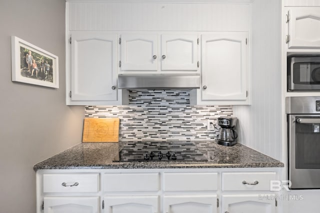 kitchen with under cabinet range hood, stainless steel appliances, white cabinetry, tasteful backsplash, and dark stone countertops