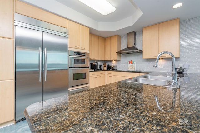 kitchen with kitchen peninsula, stainless steel appliances, wall chimney range hood, light brown cabinets, and sink