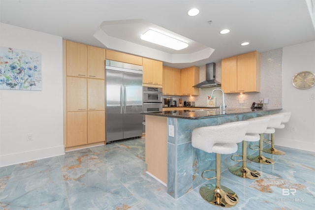 kitchen with stainless steel appliances, a raised ceiling, light brown cabinetry, and wall chimney exhaust hood