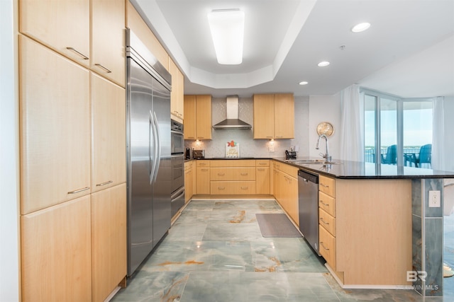 kitchen with kitchen peninsula, light brown cabinetry, wall chimney range hood, appliances with stainless steel finishes, and sink