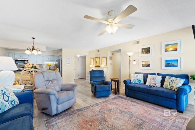 living room featuring light wood-type flooring, visible vents, a textured ceiling, and ceiling fan with notable chandelier