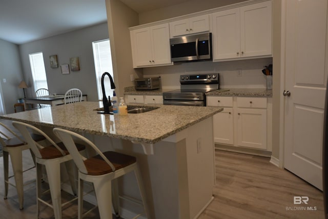 kitchen with light wood finished floors, a kitchen island with sink, a sink, appliances with stainless steel finishes, and white cabinetry