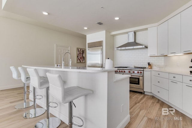 kitchen featuring light hardwood / wood-style floors, wall chimney exhaust hood, a kitchen island with sink, and premium appliances