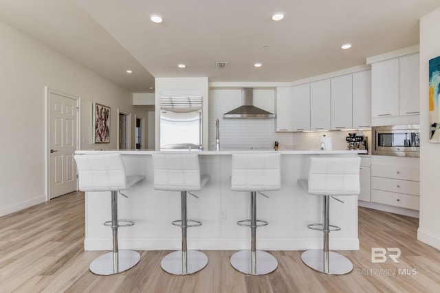 kitchen with built in appliances, wall chimney exhaust hood, white cabinetry, and a breakfast bar