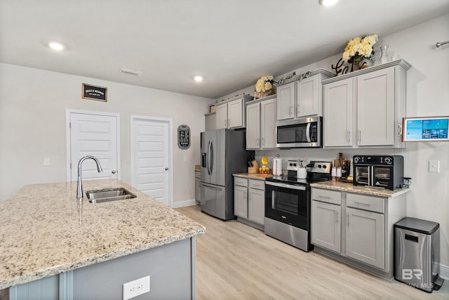 kitchen featuring gray cabinetry, stainless steel appliances, a sink, light wood-style floors, and light stone countertops