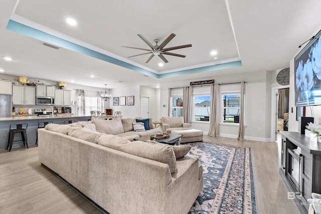 living room featuring recessed lighting, a raised ceiling, light wood-style floors, baseboards, and ceiling fan with notable chandelier