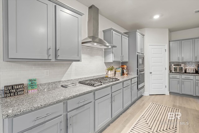 kitchen with stainless steel appliances, wall chimney range hood, gray cabinetry, light wood-type flooring, and backsplash
