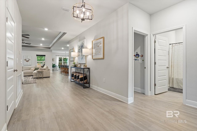 hall featuring light wood finished floors, a raised ceiling, baseboards, a chandelier, and recessed lighting
