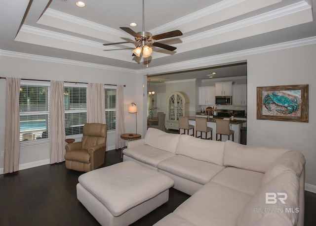 living area with ceiling fan, baseboards, dark wood-type flooring, a tray ceiling, and ornamental molding