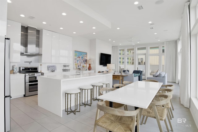 kitchen with appliances with stainless steel finishes, white cabinets, a kitchen bar, a kitchen island with sink, and wall chimney exhaust hood