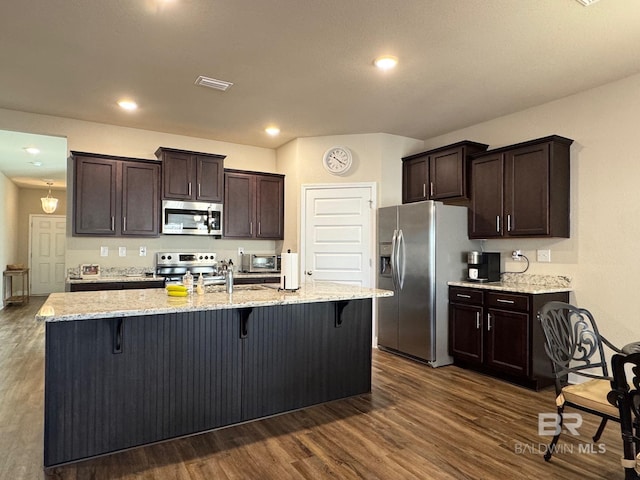 kitchen with appliances with stainless steel finishes, a kitchen breakfast bar, dark brown cabinets, a kitchen island with sink, and dark hardwood / wood-style floors