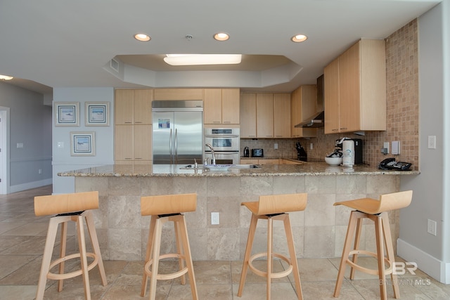 kitchen with kitchen peninsula, stainless steel appliances, light brown cabinetry, and wall chimney exhaust hood