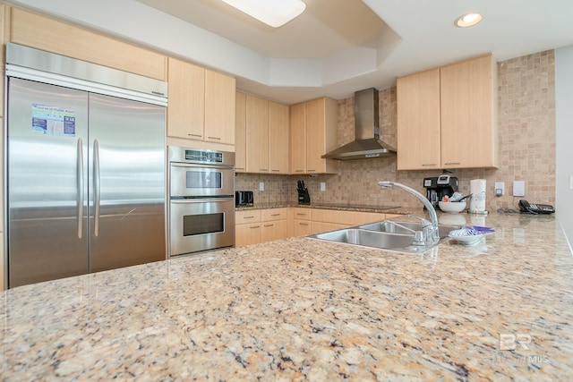 kitchen featuring appliances with stainless steel finishes, wall chimney exhaust hood, light stone counters, light brown cabinetry, and tasteful backsplash