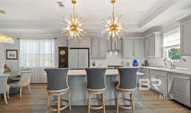 kitchen with an inviting chandelier, a tray ceiling, gray cabinets, wall chimney range hood, and stainless steel dishwasher