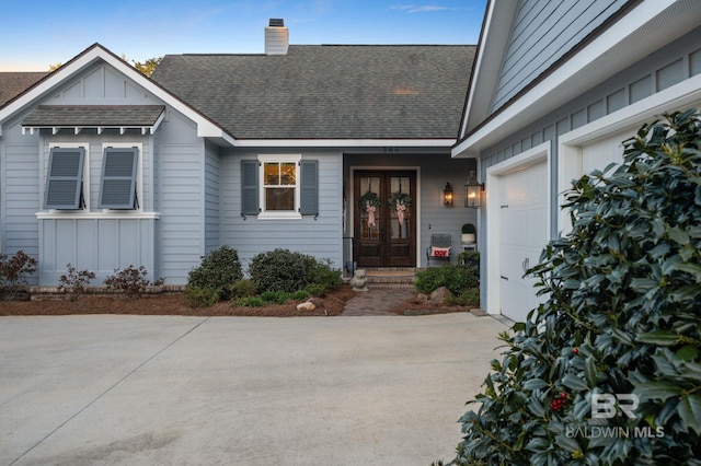 property entrance with french doors, roof with shingles, board and batten siding, concrete driveway, and a chimney