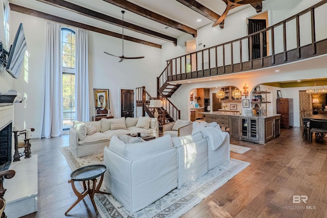 living room with a brick fireplace, hardwood / wood-style floors, beamed ceiling, a towering ceiling, and ceiling fan with notable chandelier