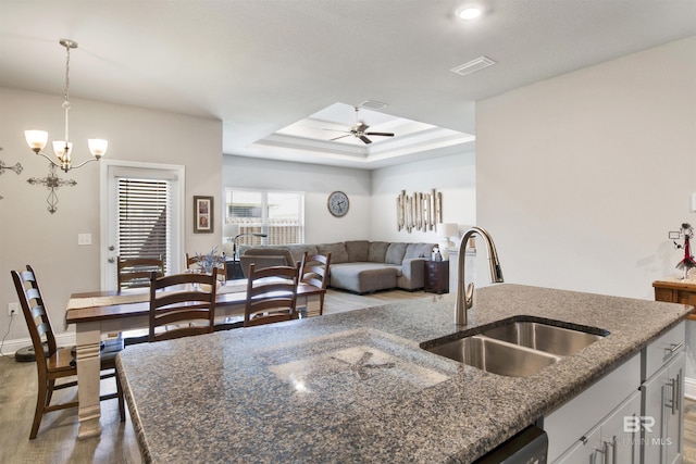 kitchen featuring visible vents, ceiling fan with notable chandelier, a sink, a tray ceiling, and dark stone countertops