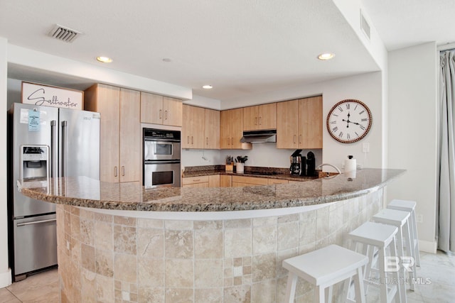 kitchen with appliances with stainless steel finishes, light brown cabinets, dark stone countertops, a peninsula, and under cabinet range hood