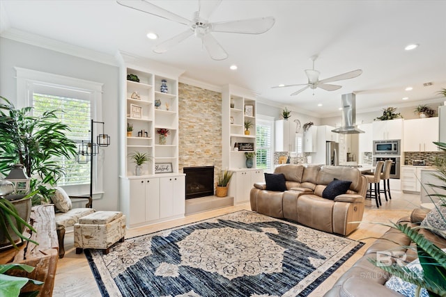 living room with built in features, a stone fireplace, crown molding, and ceiling fan