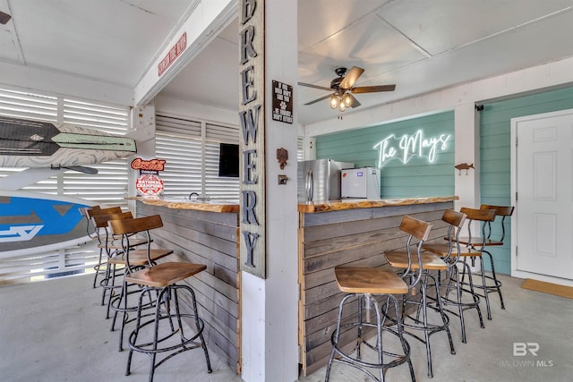 bar with concrete flooring, ceiling fan, freestanding refrigerator, and wooden walls