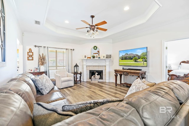 living room featuring light hardwood / wood-style flooring, crown molding, a raised ceiling, a tiled fireplace, and ceiling fan