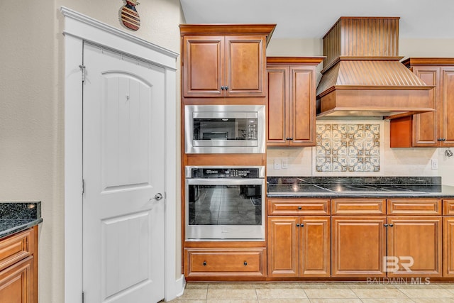 kitchen featuring custom range hood, tasteful backsplash, light tile patterned floors, dark stone counters, and stainless steel appliances