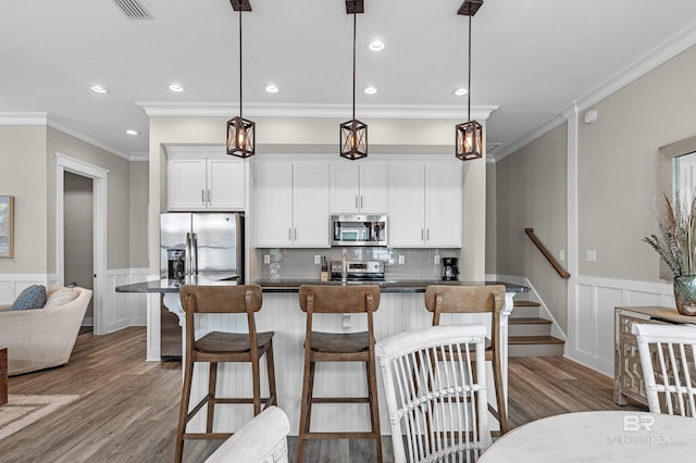 kitchen featuring dark countertops, an island with sink, decorative light fixtures, stainless steel appliances, and white cabinetry