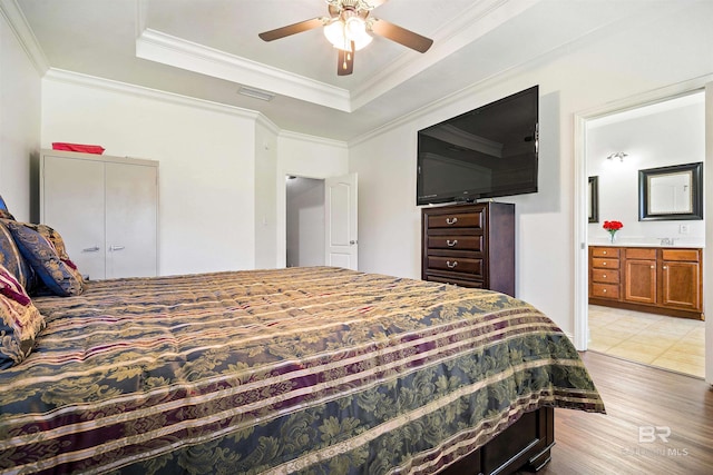 bedroom with a tray ceiling, light wood-type flooring, ensuite bath, crown molding, and a ceiling fan