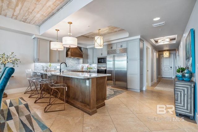 kitchen featuring light stone counters, a peninsula, decorative backsplash, built in appliances, and custom exhaust hood