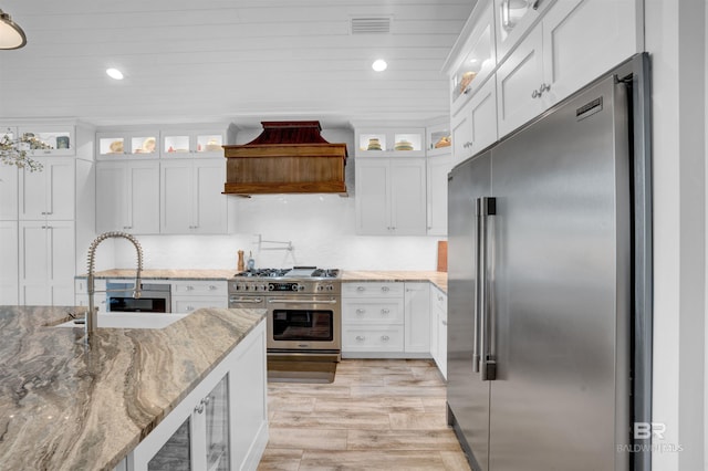 kitchen with light stone counters, premium appliances, white cabinets, light wood-type flooring, and premium range hood