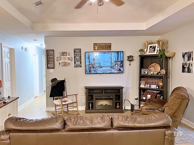 living area with a ceiling fan, light tile patterned floors, baseboards, a tray ceiling, and a glass covered fireplace