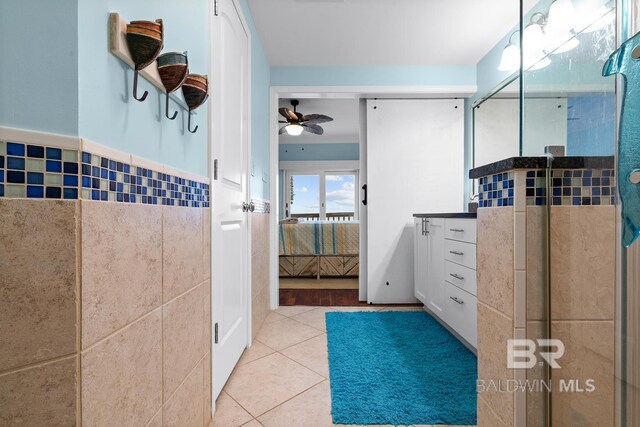 ensuite bathroom with tile walls, tile patterned floors, a ceiling fan, a shower with shower door, and wainscoting