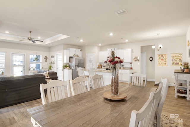 dining area with a tray ceiling, ceiling fan with notable chandelier, and light wood-type flooring