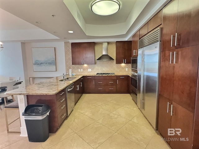 kitchen with a sink, a peninsula, wall chimney exhaust hood, decorative backsplash, and built in appliances