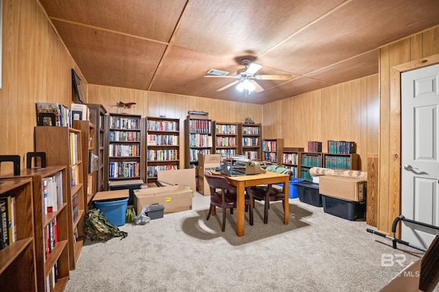 carpeted home office with bookshelves, ceiling fan, and wooden walls