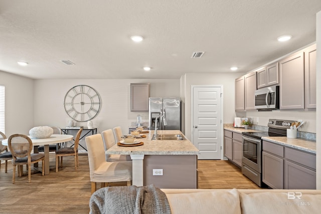 kitchen with gray cabinetry, sink, stainless steel appliances, a kitchen island with sink, and light wood-type flooring