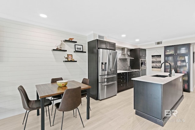 kitchen with sink, wall chimney exhaust hood, light hardwood / wood-style floors, an island with sink, and appliances with stainless steel finishes