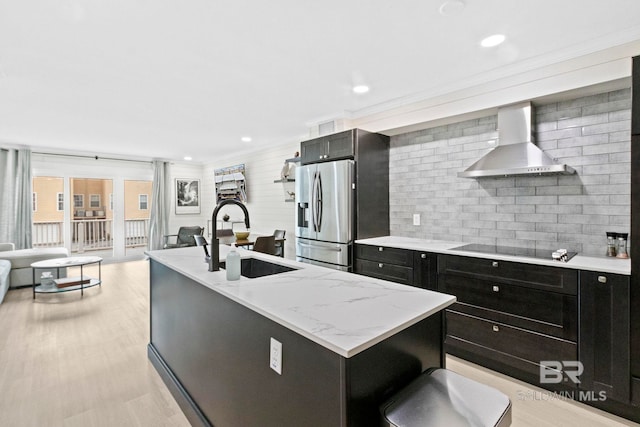 kitchen featuring an island with sink, stainless steel fridge, black electric cooktop, sink, and wall chimney range hood