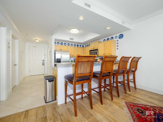 kitchen with light brown cabinetry, a kitchen breakfast bar, ornamental molding, stainless steel appliances, and light hardwood / wood-style flooring