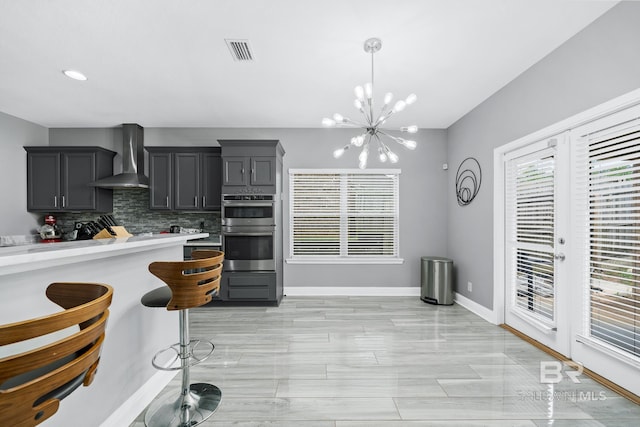 kitchen featuring backsplash, wall chimney exhaust hood, a notable chandelier, decorative light fixtures, and stainless steel double oven