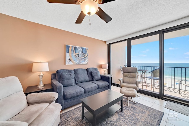 tiled living room with expansive windows, a water view, a view of the beach, ceiling fan, and a textured ceiling